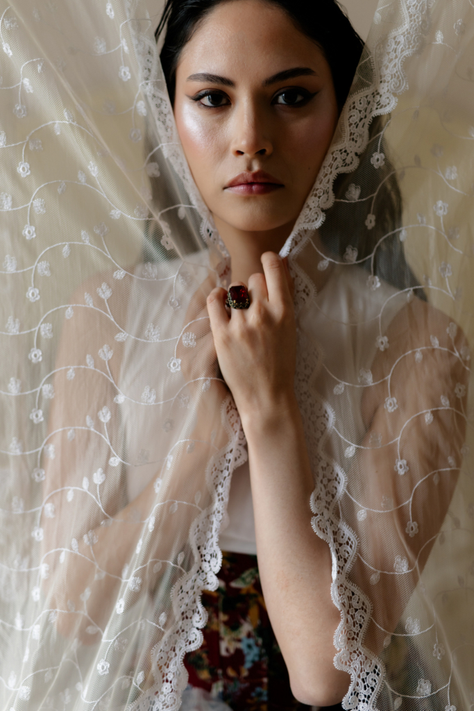 Close-up portrait of a woman framed by a delicate lace veil, looking directly at the camera with a calm expression.
Soft natural light highlights the intricate fabric details and the deep red ring on her hand.