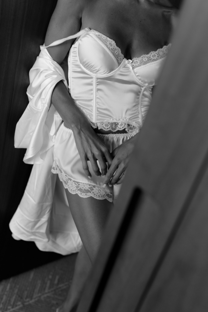 Black-and-white close-up of a woman wearing satin bridal lingerie with lace trim, partially draped in a white robe.
Soft shadows and cropped framing create an intimate, editorial-style portrait focused on texture and form.