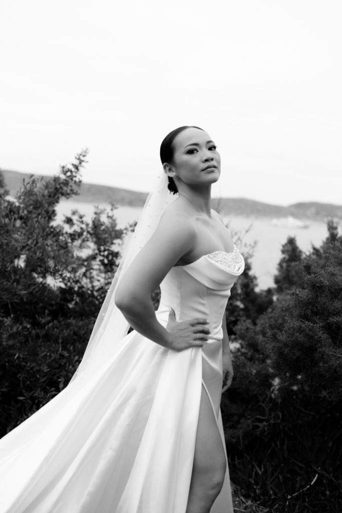 A bride in a flowing satin gown stands against a soft coastal backdrop, photographed in elegant black and white.
Her poised expression, sweeping veil, and sculptural silhouette give the portrait a timeless cinematic quality.