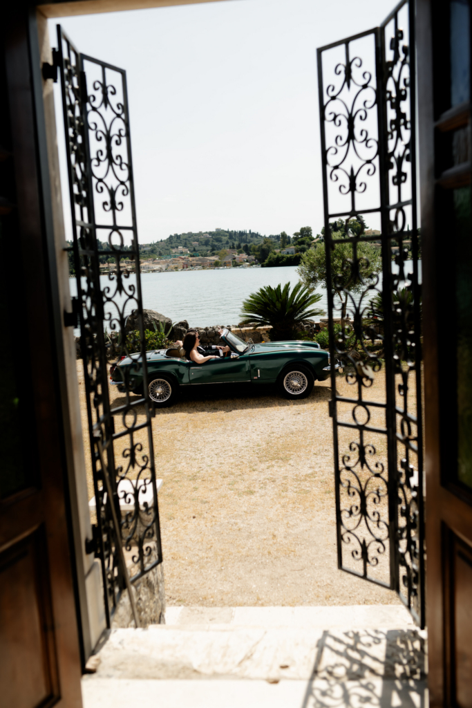 View through open wrought-iron doors toward a vintage green convertible parked beside the water on a sunny day.
The scene combines Mediterranean architecture, still water, and classic car elegance in a cinematic frame.