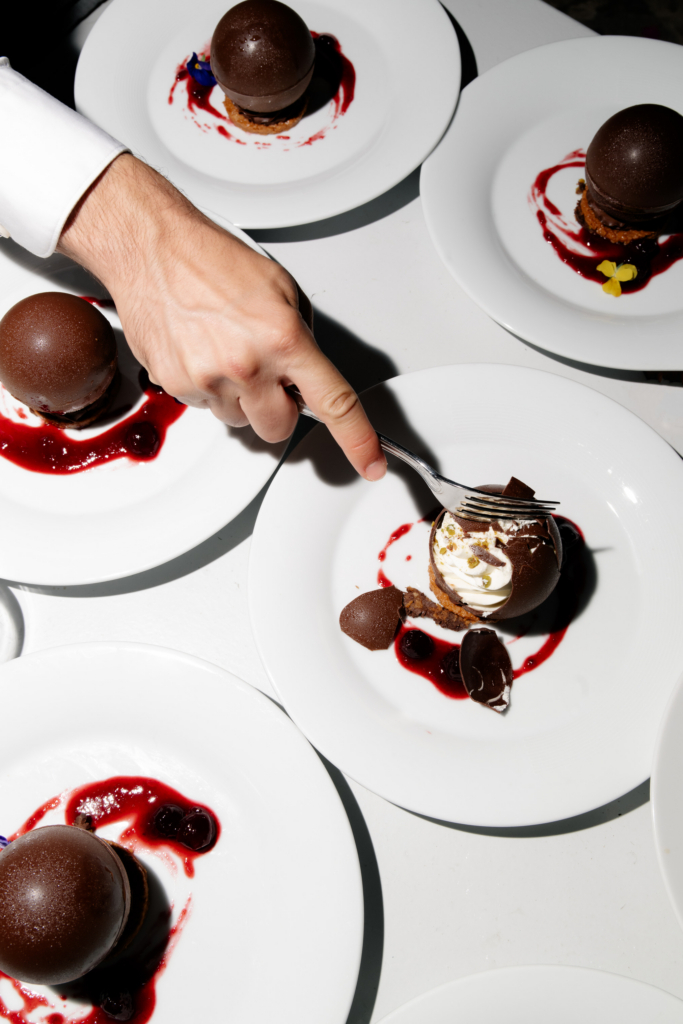 Overhead view of plated chocolate sphere desserts drizzled with red berry sauce as a hand breaks one open with a fork.
Strong sunlight and graphic shadows create a dramatic, editorial-style food photograph.