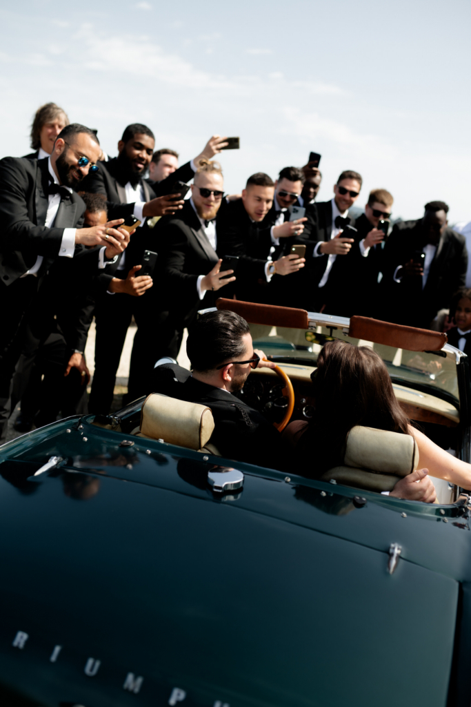 Guests in black tuxedos gather around a vintage convertible, holding up their phones to photograph the couple seated inside the car.
The candid moment captures the excitement and energy of a wedding celebration under bright daylight.