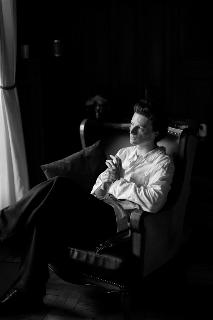 Black-and-white portrait of a person seated in a leather armchair beside a softly lit window, wearing a loose white shirt and dark trousers.
The low light and deep shadows create a quiet, introspective atmosphere.