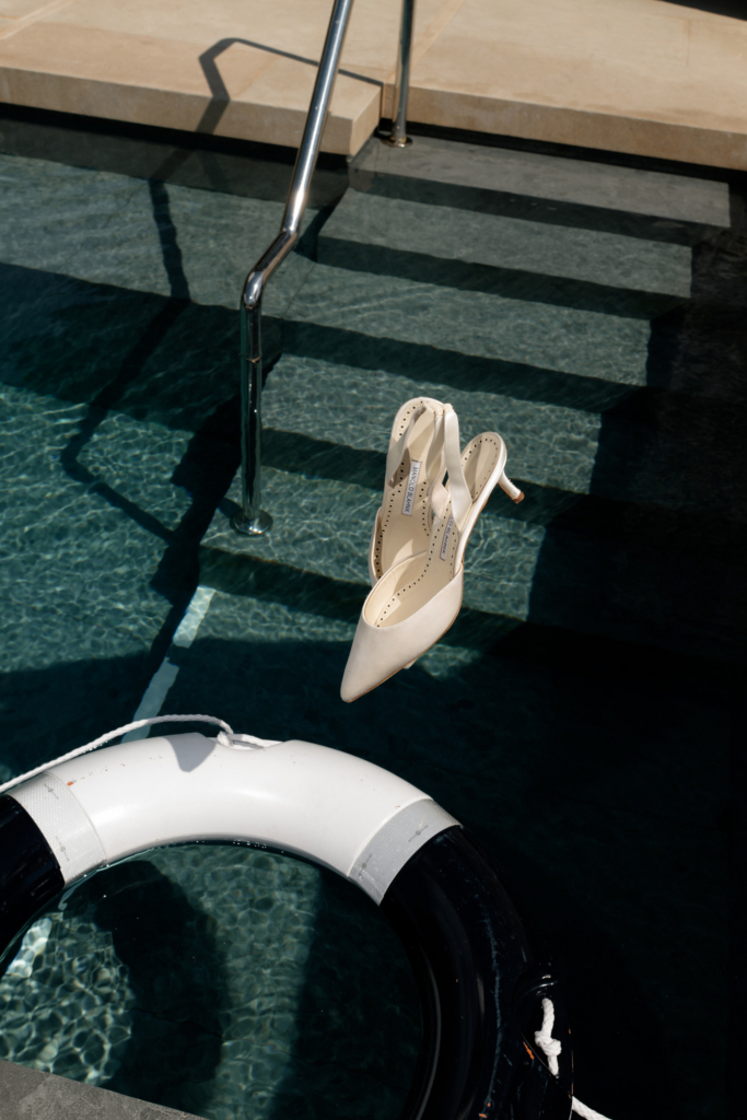 A pair of white satin bridal heels balanced above a dark swimming pool beside a floating life ring and sunlit steps.
Strong shadows and reflective water create a graphic, cinematic composition with a Mediterranean summer mood.