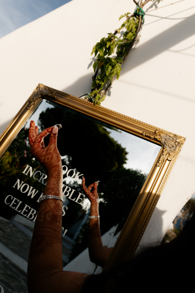 A hand decorated with intricate henna designs holds a ring in front of an ornate gold-framed mirror outdoors at sunset.
Warm light, reflections, and ceremonial details create an intimate and elegant composition.