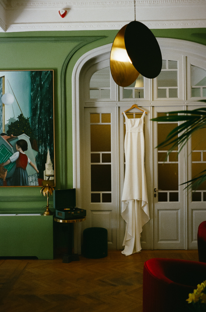 A minimalist white wedding dress hangs from tall paneled doors inside an elegant room with green walls and warm ambient light.
Vintage decor, framed artwork, and soft shadows create a quiet, cinematic atmosphere before the ceremony.