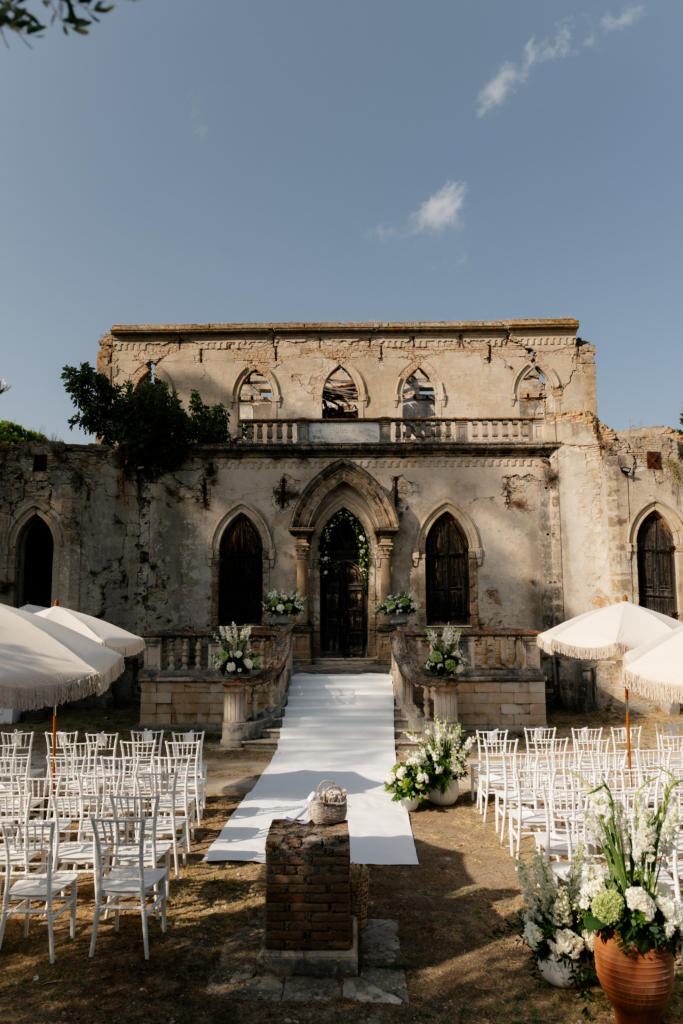 An outdoor wedding ceremony setup sits in front of a weathered historic stone building with Gothic-style arches and crumbling textures.
The symmetry of the white aisle, floral arrangements, and aged architecture creates a cinematic contrast between elegance and decay.