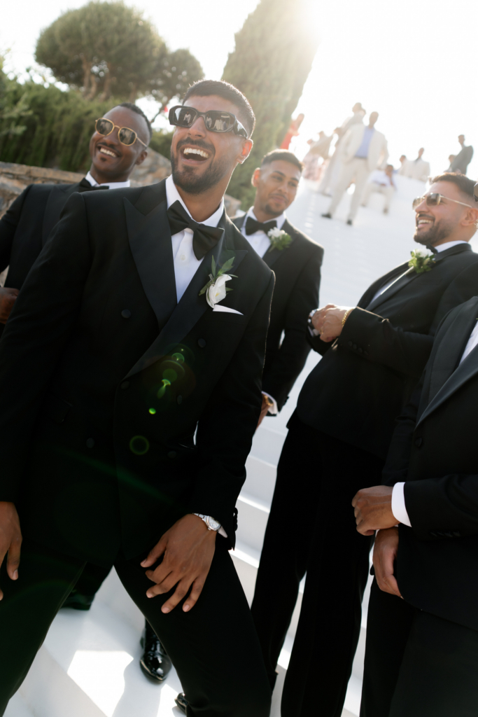 Group of groomsmen in black tuxedos and sunglasses laughing together on sunlit white steps during an outdoor wedding celebration.
Bright backlight and lens flare add energy and a candid, cinematic atmosphere to the scene.