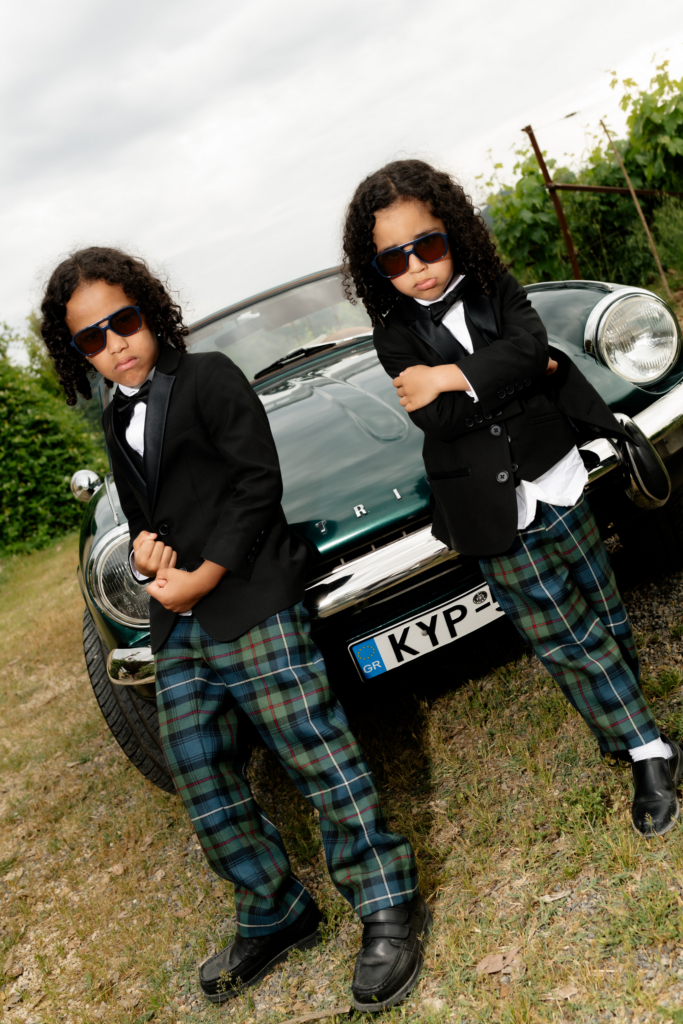 Two children wearing tuxedo jackets, plaid trousers, and sunglasses pose confidently in front of a vintage green convertible.
The playful portrait combines formal wedding style with a cinematic, humorous attitude.