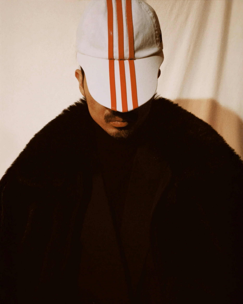 A close-up portrait of a man wearing a white cap with bold red stripes that obscures his eyes. He is dressed in a dark, textured coat, with warm light casting soft shadows across his face.