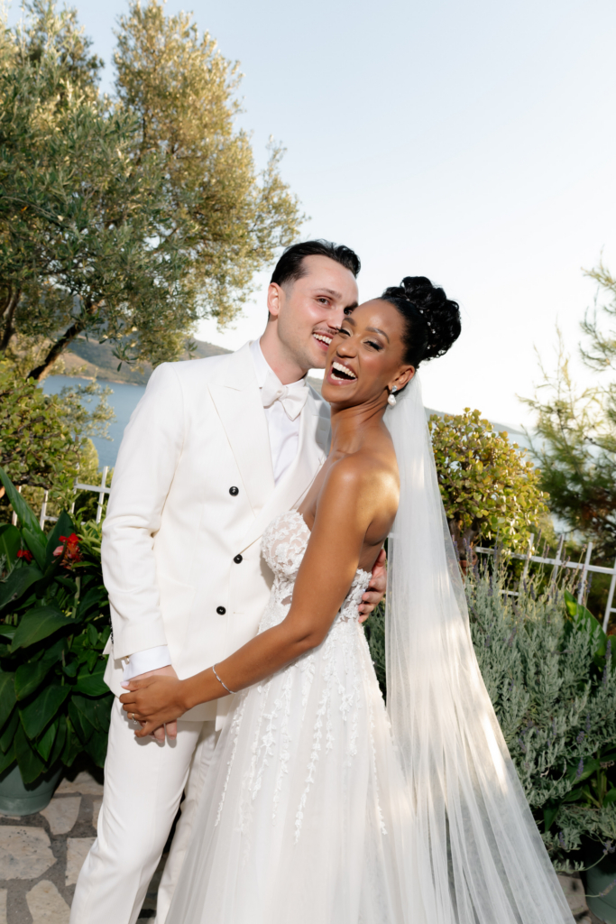 Bride and groom posing together in a garden overlooking the sea, both dressed in white formalwear.
The portrait captures a joyful, relaxed moment during an outdoor wedding celebration.