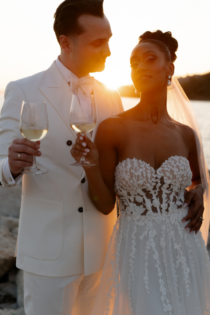 Bride and groom holding glasses of white wine at sunset by the water, illuminated by warm golden light.
The backlit portrait creates an intimate and cinematic atmosphere.