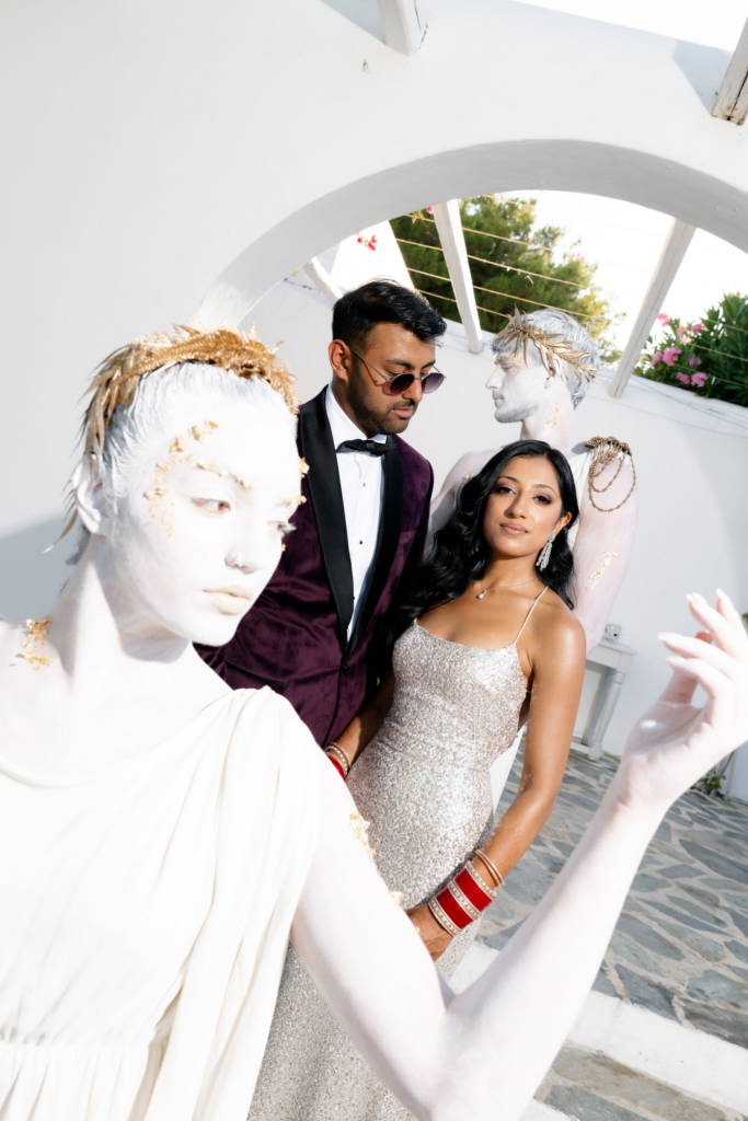 A formally dressed couple stands among white-painted performance artists wearing gold laurel crowns in a sunlit courtyard.
The image blends fashion, sculpture, and ceremony with a surreal, editorial atmosphere.