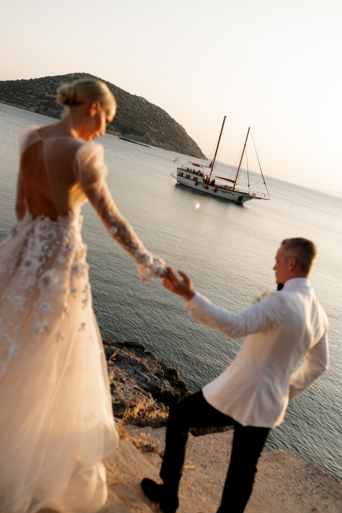 Bride and groom holding hands while walking along a rocky seaside cliff at sunset, with a sailboat drifting across calm water in the background.
The warm evening light and tilted composition create a cinematic, dreamlike feeling.