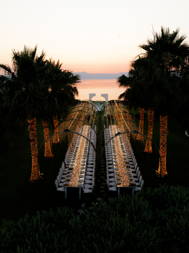 A long candlelit reception table stretches toward the sea at sunset, framed by glowing palm trees and a soft pastel horizon.
The symmetry, warm lighting, and coastal backdrop give the scene a cinematic destination wedding atmosphere.
