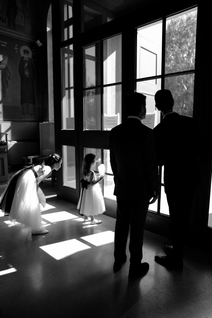 Inside a softly lit church, two flower girls and two suited guests stand silhouetted against towering glass doors as sunlight spills across the floor.
The black-and-white composition and quiet gestures create a cinematic moment that feels both intimate and observational.