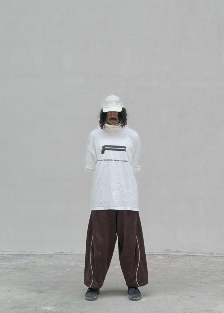 Photographer Jay Zoo stands against a plain white wall in an oversized white T-shirt, wide brown trousers, and a white cap. His posture is still and centered, with a minimal, understated presence.