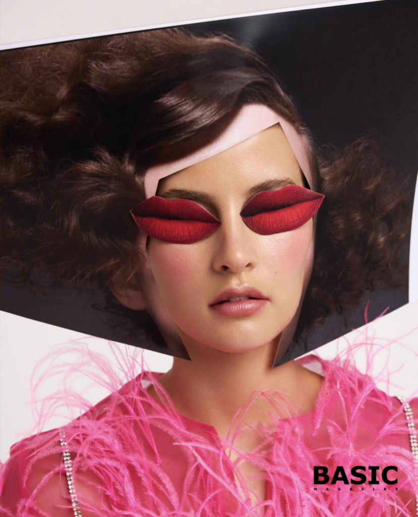 A surreal fashion beauty portrait with a strong editorial concept: a model framed inside a printed image of herself, with sculptural red lips placed over the eyes like a visual mask. The styling is bold, graphic, and playful, while the pink feathered garment adds softness against the sharp composition.