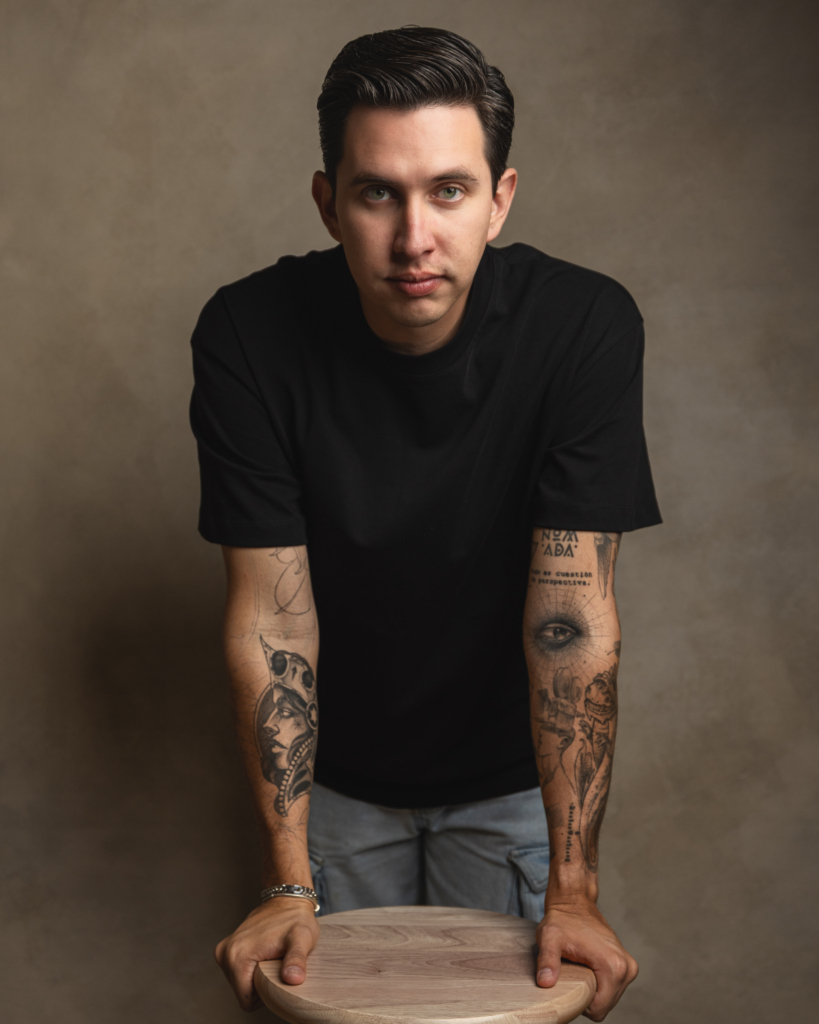 A portrait of photographer Miguel Angel “Demo” standing behind a wooden stool, looking directly into the camera. He wears a plain black t-shirt, and his tattooed arms are visible against a soft neutral studio background.