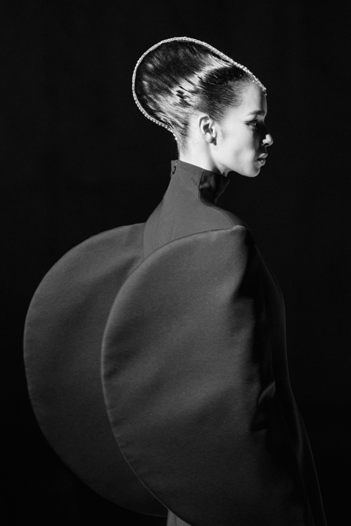 A black-and-white portrait of a model in a sculptural garment with exaggerated rounded shoulders. Her profile and sleek, structured hair create a sharp, graphic silhouette against a dark background.