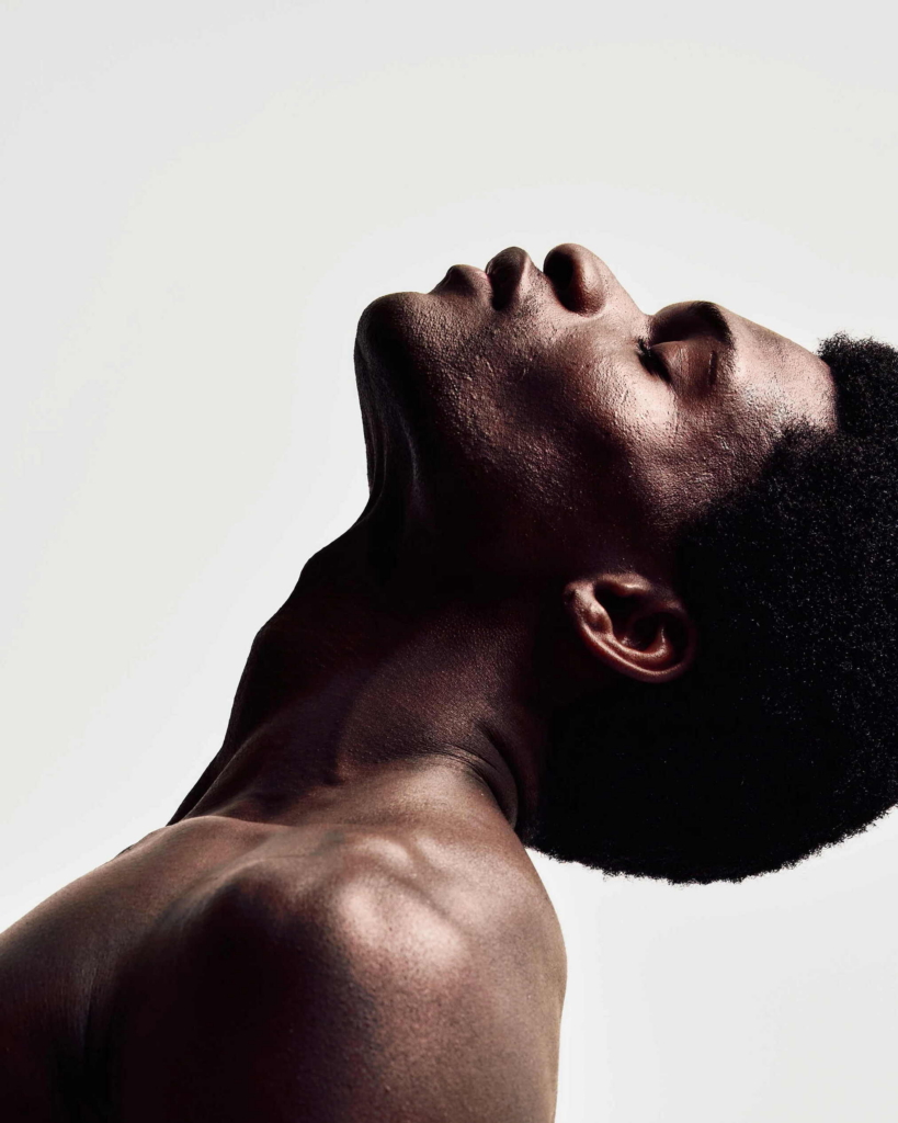 A close-up portrait of a shirtless model with his head tilted back and eyes closed. Strong directional light emphasizes the contours of his face, neck, and shoulder against a plain background.