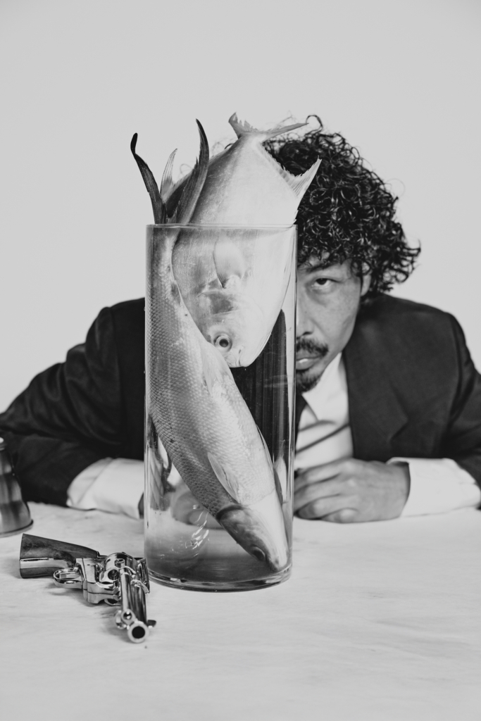 A striking black-and-white editorial series that blurs the line between still life and portraiture. The surreal composition, cinematic styling, and carefully placed objects create a frame that feels both theatrical and unsettling, while the subject’s presence adds a sharp sense of character, mystery, and visual tension.