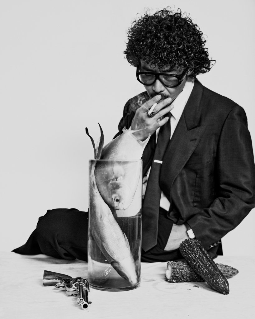 A striking black-and-white editorial series that blurs the line between still life and portraiture. The surreal composition, cinematic styling, and carefully placed objects create a frame that feels both theatrical and unsettling, while the subject’s presence adds a sharp sense of character, mystery, and visual tension.