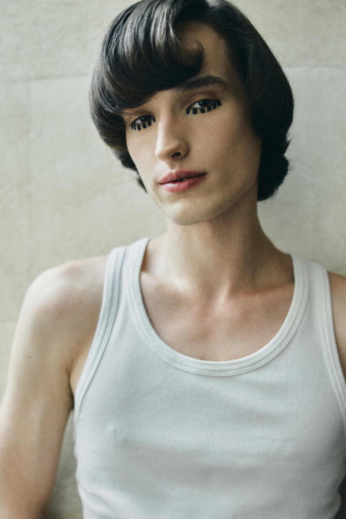 A soft portrait of a model in a white tank top, with short sculpted hair and graphic, stylized eyelashes. The image feels quiet and intimate, with a subtle focus on texture and detail.