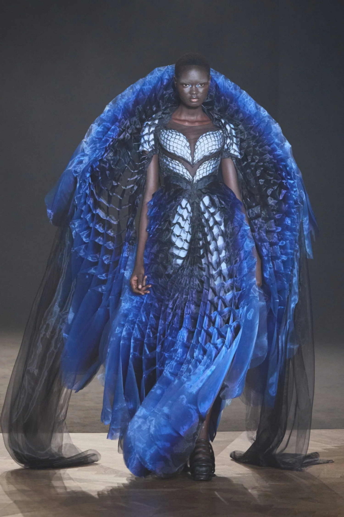 A runway image of a model wearing a dramatic blue and black sculptural gown with layered, wing-like volume. The silhouette is bold and theatrical, with the garment dominating the frame as the model walks forward.