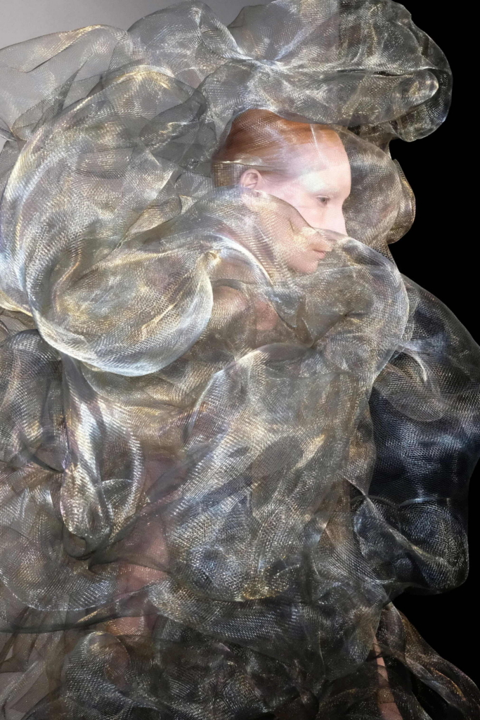 A portrait of a model partially wrapped in layers of shimmering, translucent silver fabric. The material billows around the face and body, creating a sculptural, otherworldly silhouette.