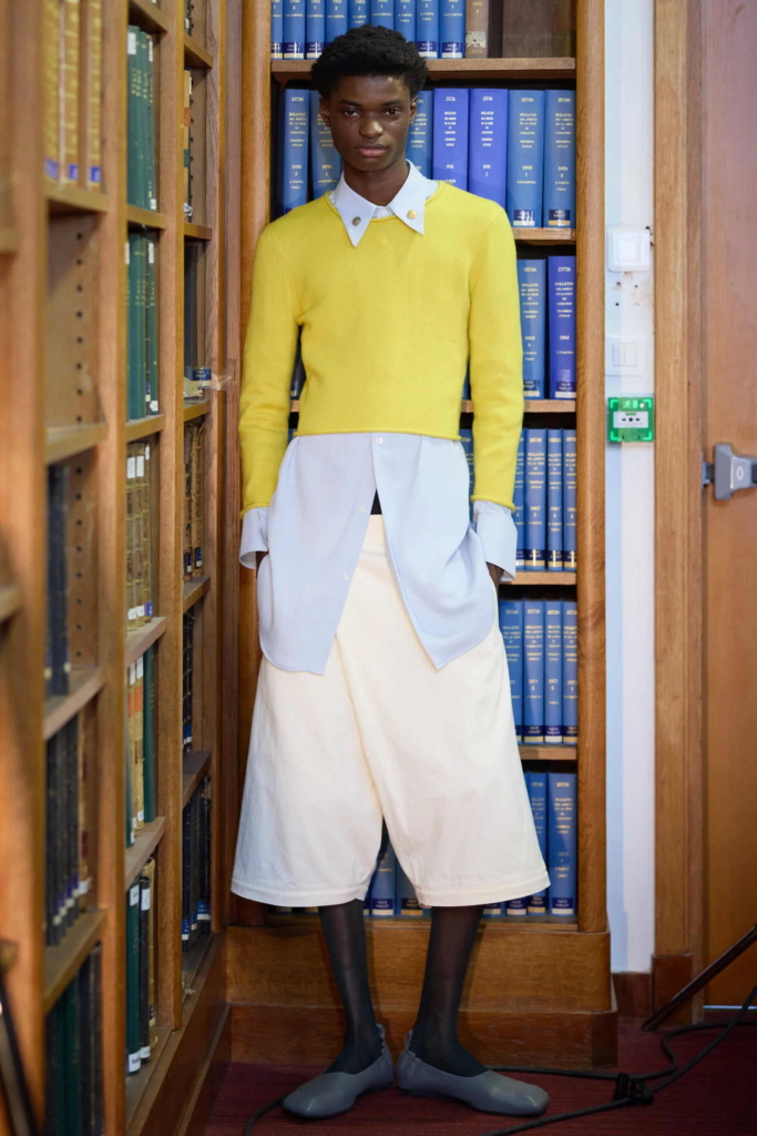 A fashion portrait of a model standing between library shelves in layered yellow, pale blue, and cream clothing. The setting adds a quiet, unexpected contrast to the clean, structured styling.