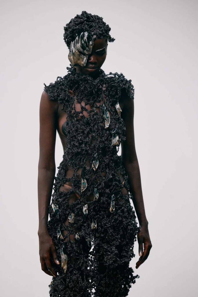 A portrait of a model wearing a dark, sculptural garment with metallic elements partially covering one eye. The look feels futuristic and organic at once, with a strong sense of texture and presence.