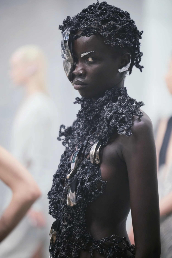 A portrait of a model wearing a dark, sculptural garment with metallic elements partially covering one eye. The look feels futuristic and organic at once, with a strong sense of texture and presence.