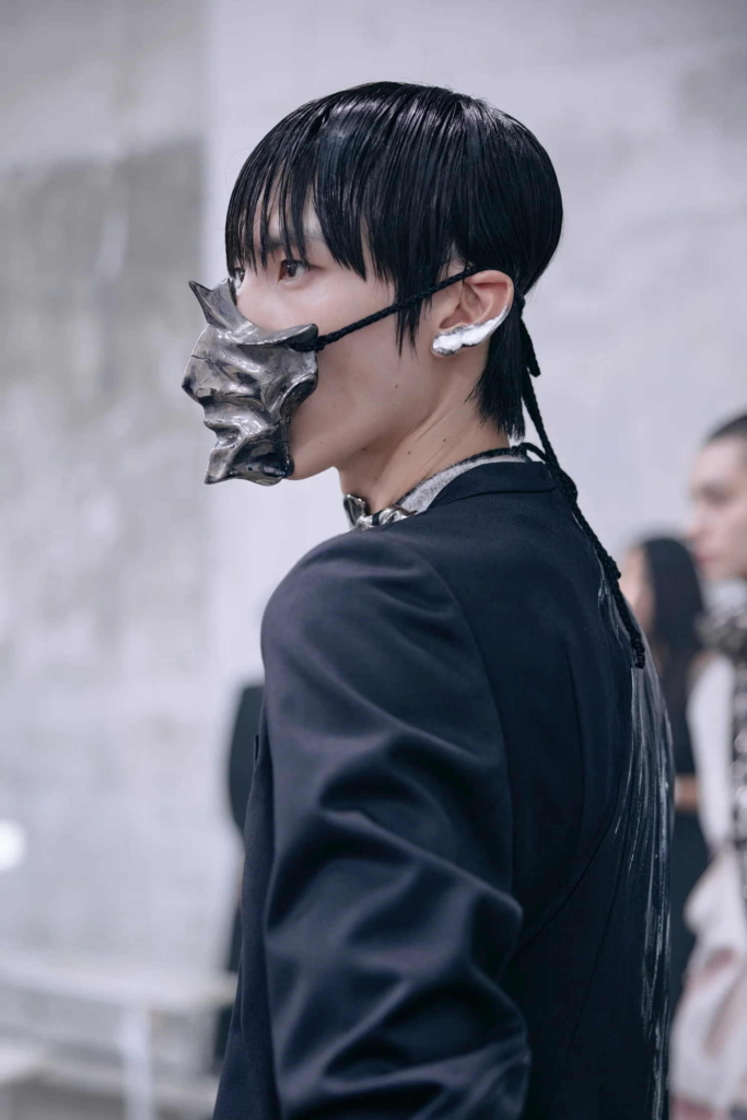 A backstage fashion portrait of a model in a black tailored jacket, wearing a sculptural metallic face piece. The image captures a moment of stillness amid the movement of a runway environment.