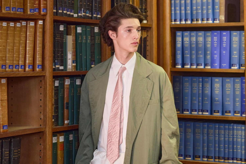 A fashion portrait of a model in a pale green jacket, white shirt, and soft pink tie, standing in front of tall library shelves. The image balances formal styling with a slightly offbeat, editorial mood.