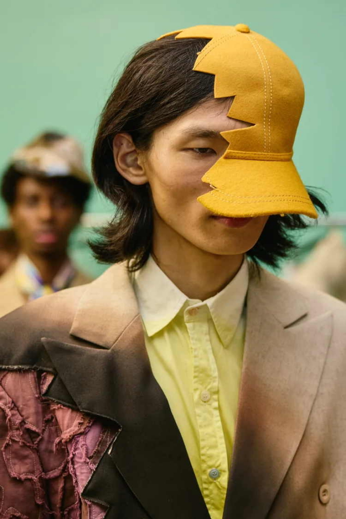 A close-up backstage fashion portrait of a model wearing a sculptural yellow cap that covers half the face. The look combines soft tailoring, sharp styling, and an offbeat sense of design.