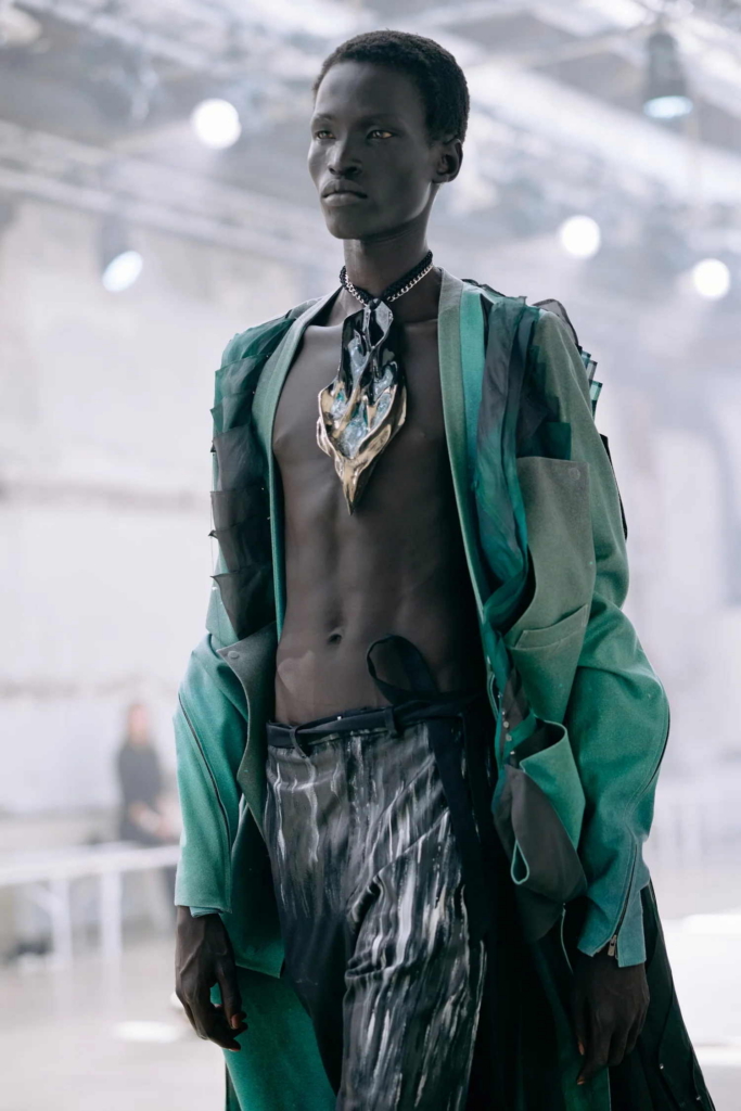 A backstage fashion portrait of a model wearing layered green and black styling with a sculptural metallic chest piece. The image feels futuristic and composed, with a quiet intensity against the soft runway background.
