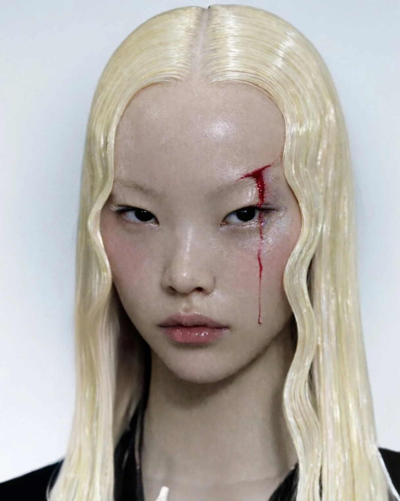 An AI-generated close-up portrait of a woman with long platinum-blonde hair and a calm, direct expression. A thin red cut above one eye runs down her face, creating a sharp contrast against the otherwise clean, polished image.