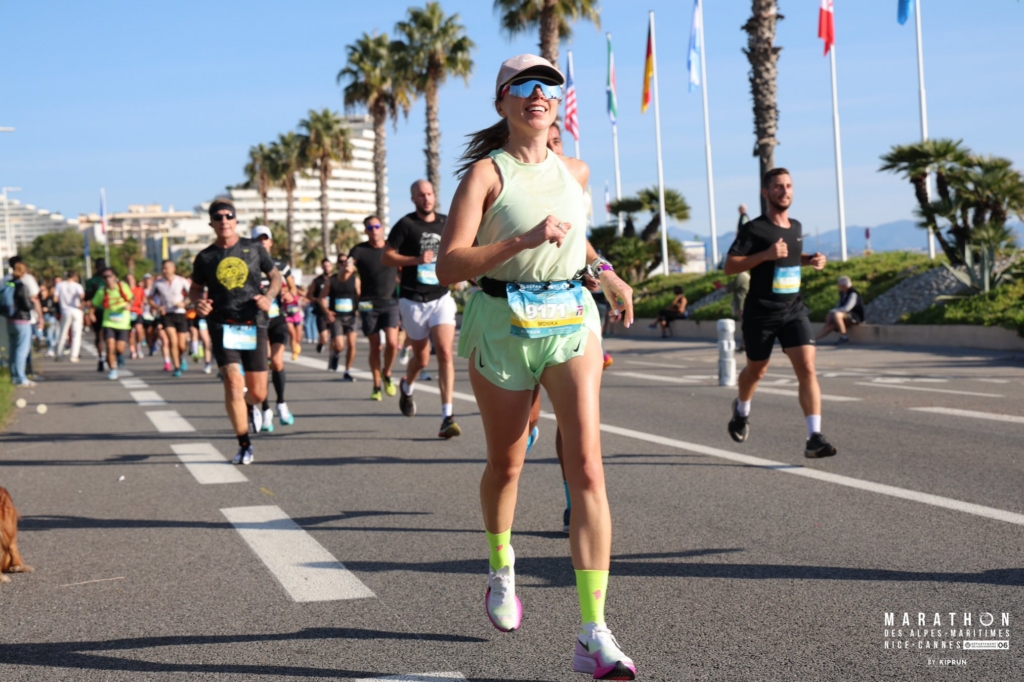 Monika runs in a marathon along a palm-lined road on a sunny day, surrounded by other runners. She wears a light athletic outfit, sunglasses, and a race bib, moving with focus and energy. The scene captures motion and determination in a bright, open setting.