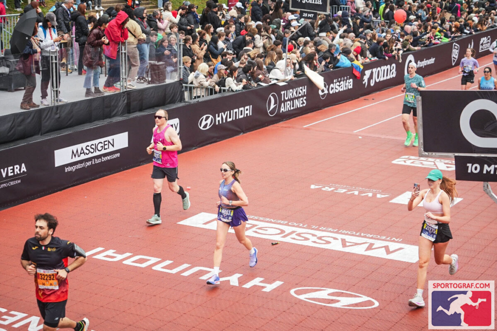 Runners approach the finish area of a marathon, with a large crowd cheering from behind barriers. Monika is among them, mid-stride, focused as she moves toward the end of the race. The scene feels energetic and celebratory, with spectators adding to the intensity of the moment.