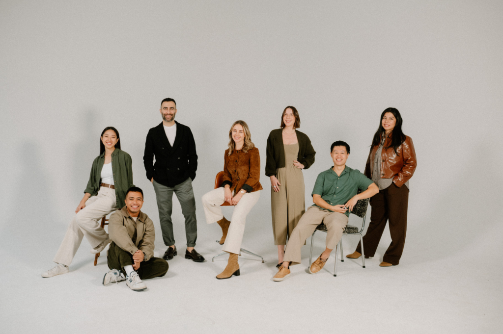 A diverse group of seven adults poses together in a bright, minimalist studio with a clean, neutral background. Some are standing while others sit casually on chairs or the floor, dressed in coordinated earthy tones and relaxed styles. The group appears natural and approachable—this is Monika’s team.