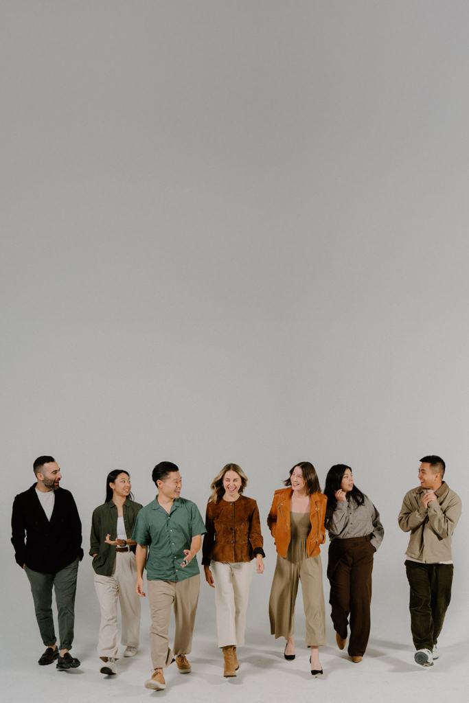 A group of seven adults walks side by side in a minimalist studio with a clean, light background, engaged in conversation and laughter. Their relaxed body language and natural expressions suggest familiarity and ease, with coordinated neutral and earthy-toned outfits. The scene captures a candid, dynamic moment—Monika’s team moving together with a sense of connection.