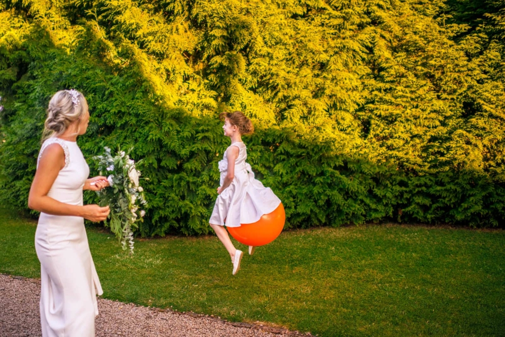 A bride stands on the left holding a bouquet, looking toward a young girl bouncing on an orange ball. Behind them is a dense green hedge in warm sunlight.