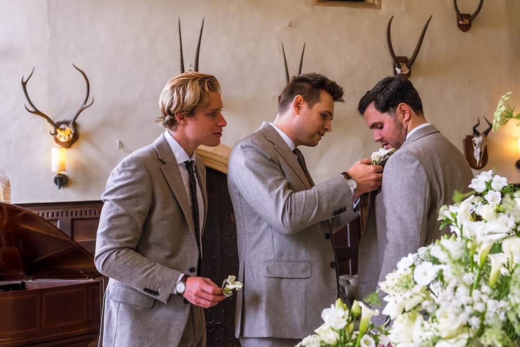 Three men in light grey suits stand together in a softly lit room, preparing for a wedding. One carefully pins a white boutonniere onto another man’s lapel while the third waits beside them holding an extra flower.
The background features cream walls, mounted antlers, warm wall lights, and a large arrangement of white flowers in the foreground, giving the scene a calm, elegant atmosphere.