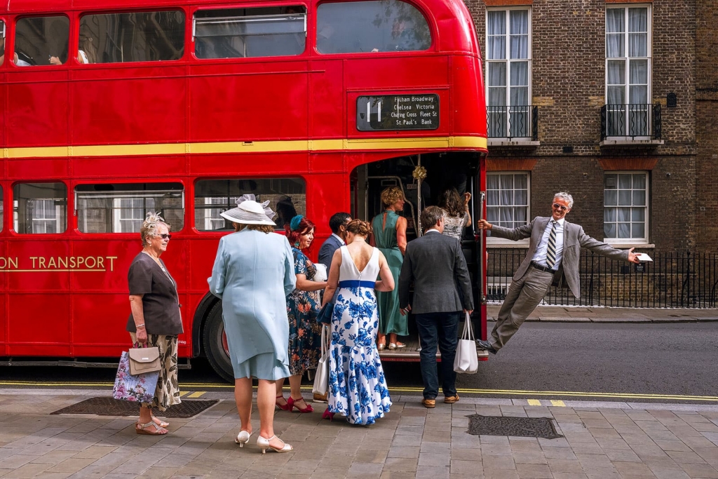 A bright red double-decker bus is parked on a city street while a group of wedding guests in formal clothes climb aboard. One man in a gray suit stands at the bus door with his arms stretched wide, smiling theatrically as if welcoming everyone inside.