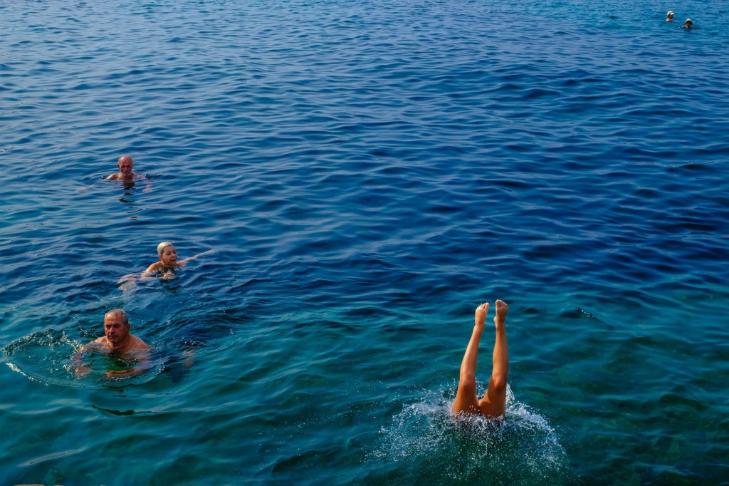 Several people swim in open blue water, spaced apart from each other. In the foreground, a person dives underwater with only their legs visible above the surface. Gentle ripples spread across the sea under bright daylight.
