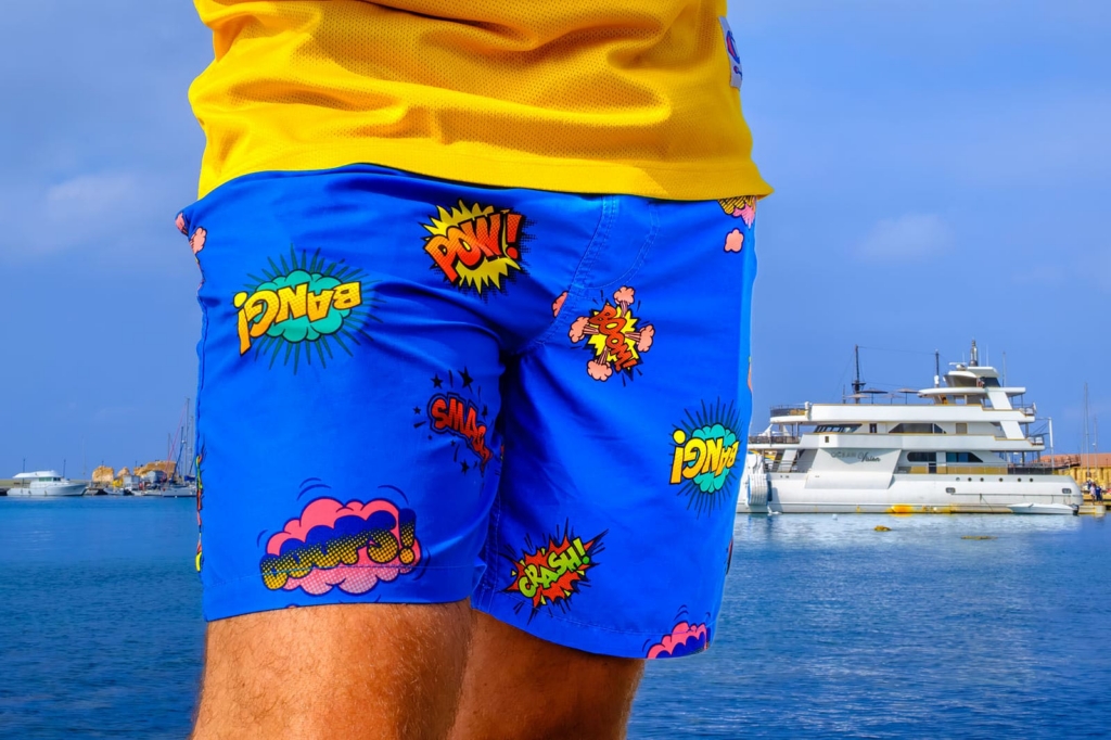 A close-up view shows the lower half of a person standing by the water, wearing bright blue swim shorts covered in colorful comic-style words and symbols. Behind them, the sea is calm and several white yachts are docked in the marina under a clear blue sky.