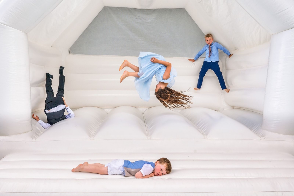 An overhead view shows four children scattered across a large white inflatable bouncy castle. One girl in a light blue dress is mid-flip in the air, while another child lies face-down at the bottom. Two boys in dress clothes are positioned at opposite sides, one standing and one upside down against the wall.