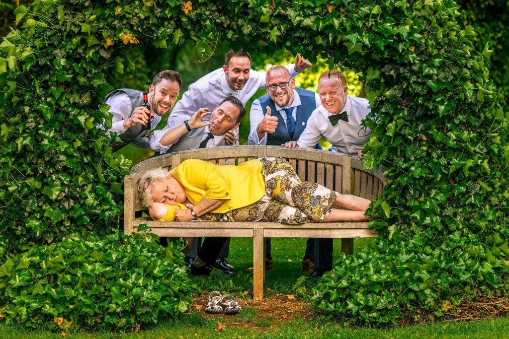 An older woman in a bright yellow jacket lies curled up on a wooden bench as if asleep, framed by a dense arch of green ivy. Behind the bench, five men in wedding clothes lean in with playful expressions, one holding a beer and another giving a thumbs-up. A pair of shoes sits on the grass beneath the bench.
