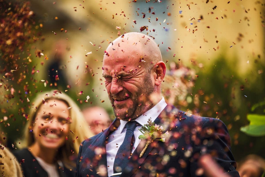 A bald man in a suit stands outdoors as colorful confetti bursts around him, his eyes tightly shut and face scrunched in reaction.
Behind him, smiling guests are slightly out of focus, creating a lively, celebratory wedding atmosphere.